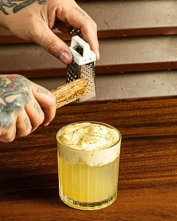 Mãos tatuadas ralando especiaria sobre um coquetel com espuma